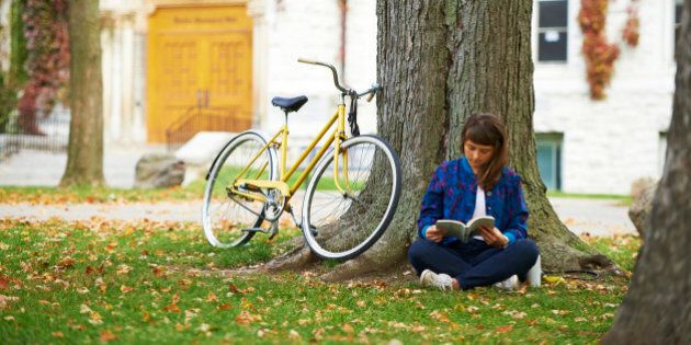 Girl reading book on university campus in fall with yellow bicycle; Kingston, Ontario, Canada