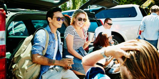 Smiling friends sitting on bumper of car tailgating in stadium parking lot before football game