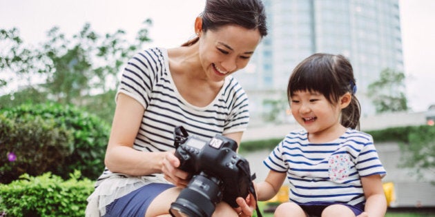 Pretty young mom showing pictures to lovely little girl from the back of the camera on the stone steps in the park in front of commercial buildings, while both smiling joyfully.