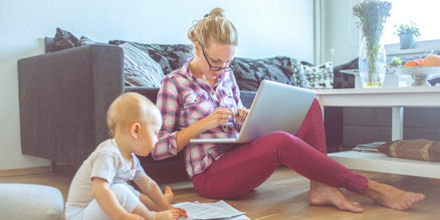 Attractive blonde woman working on a laptop computer while her barefoot baby plays around. The woman is casually dressed with long blonde hair that is swept back from her face. She looks concentrated and she is probably doing her budget or paying bills. Also, she could be a business woman that works at home. The shot is executed with available natural light, and the copy space has been left. Shallow DOF. Soft focused.