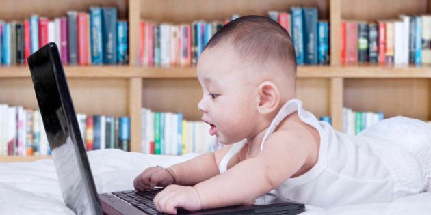 Portrait of cute baby lying on bed and using laptop computer with bookshelf background