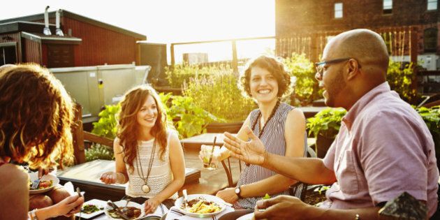 Laughing group of friends sitting together sharing dinner and wine in rooftop garden on summer evening