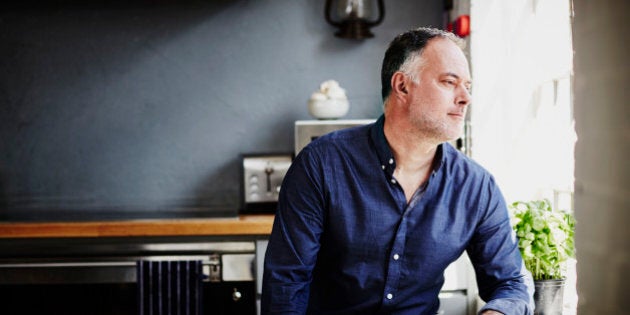 A man wearing a shirt sits in his kitchen looking out of window on a sunny day