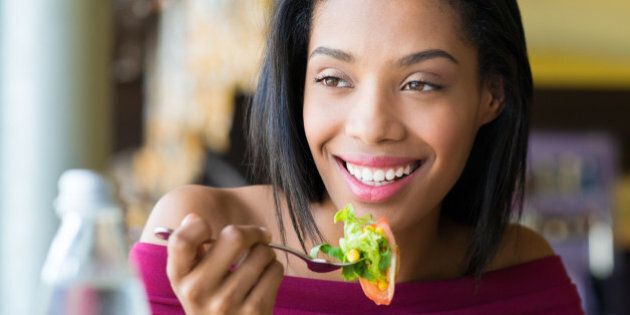 Closeup shot of young woman eating fresh salad at restaurant. Healthy african girl eating salad and looking away. Smiling young woman holding a forkful of salad. Health and diet concept. Woman ina a lunch break.