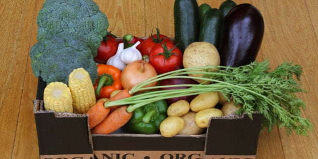 Box filled with organic vegetables, carrots, aubergine, courgettes, tomatoes, pototoes, onions, garlic, peppers, broccoli and corn on the cob, on wooden floor.