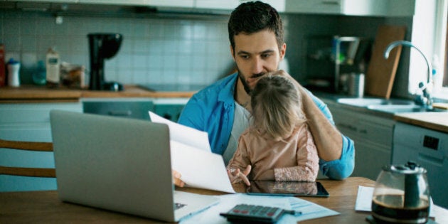 Photo of a father and daughter sitting in the kitchen,daughter is playing while dad is sitting worried and checking his bills