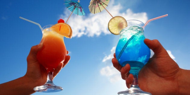 holding tropical cocktails with umbrellas, ice, fruit and straws against a blue sunny sky