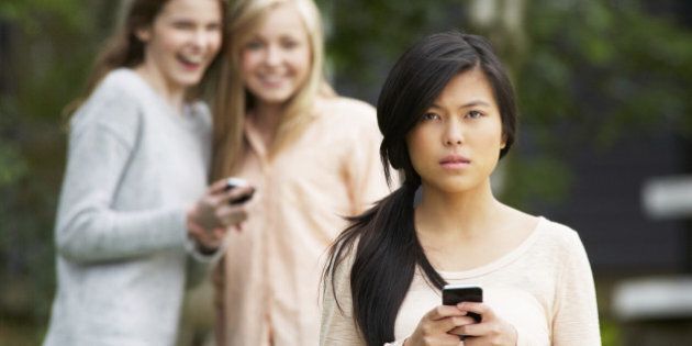 Teenage Girl Being Bullied By Text Message On Mobile Phone With Bullies In Background