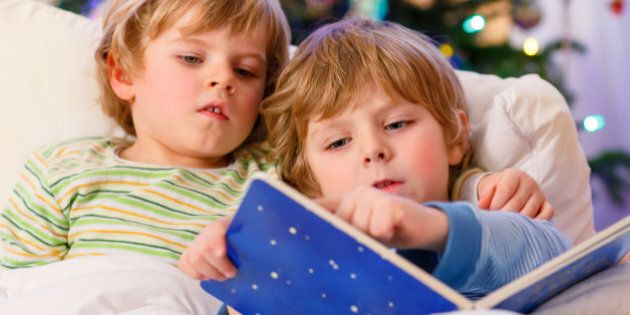 Two little blond sibling boys reading a book in bed near Christmas tree with lights and illumination. Happy family of two brothers.