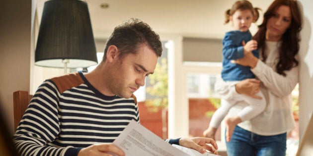 A young husband and father sits at a dining table looking through the household bills and looks worried . His wife and young daughter stand in the background looking on sympathetically .