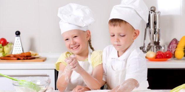 Happy little boy and girl wearing a white chefs uniform and hat cooking in the kitchen standing at the counter making a batch of biscuits and rolling the dough
