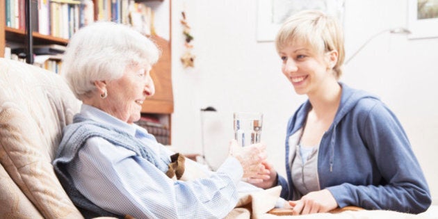 Young home caregiver giving a glass of water to senior woman, 89 years old, sitting on chair, focus on senior woman