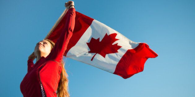 A young woman patriotically with long blond hair holds the Canadian flag above her head, as it blows in the breeze against a pristine blue sky.