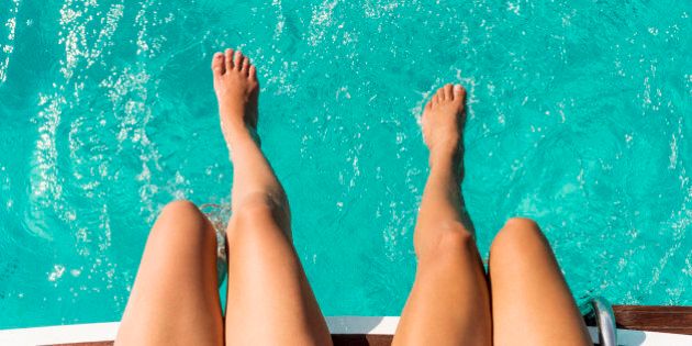 two womans legs dangeling over back of boat with there feet in clear blue ocean water