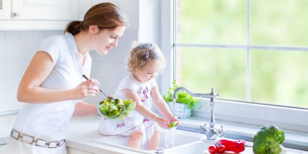 Young beautiful mother and her cute curly toddler daughter washing vegetables together in a kitchen sink getting ready to cook salad for lunch in a sunny white kitchen with a big garden view window