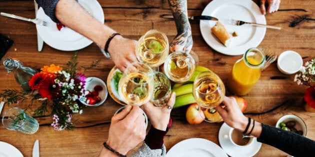 Young people sitting in a bright cafÃ© are having a toast with glasses of white wine during dinner. The glasses are seen from bird's eye view while there is food and flowers on the table.