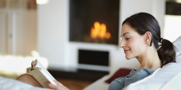 A young woman is lying on a white couch reading a book. In the backround there is a fireplace.