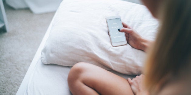 Young woman sitting at the bed in her bedroom, holding smartphone with alarm application on the screen.