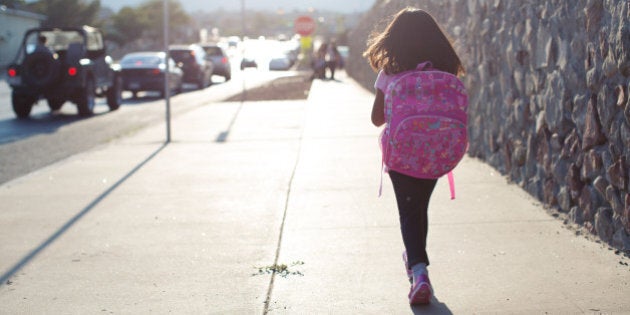 A young girl walks to school along a busy street wearing her backpack.