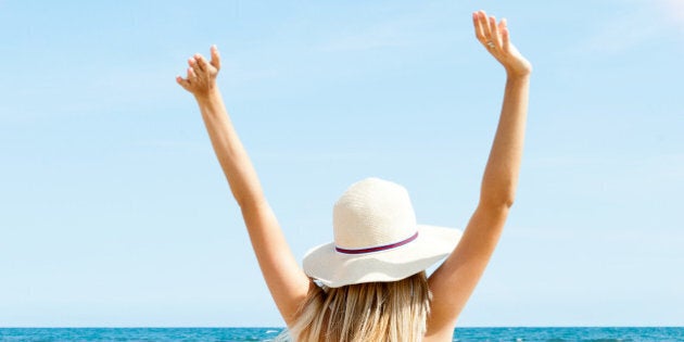 Portrait of young woman on the beach near the sea sitting with hands up wearing sophistical dress and hat looking at rainbow. Photo from behind