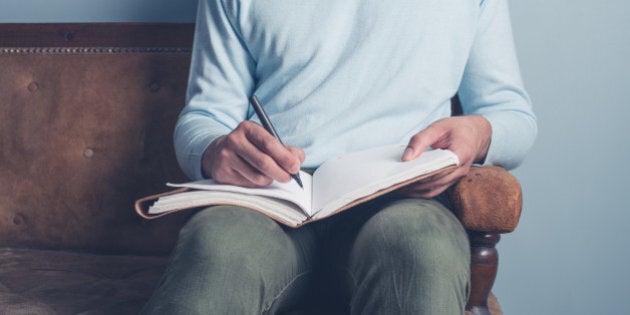 A young man is sitting on an old sofa and is writing in a notebook