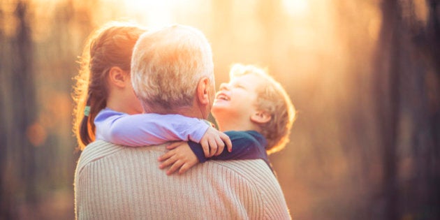 Picture of a grandfather playing with his two grand children a five year old granddaughter and three year old grandson in the park. The old man is wearing a khaki sweater and a blue shirt with long sleeves. The little girl is wearing a pink blouse and pink sweatpants and boy is wearing blue blouse. They are smiling and having fun. The lens flare is behind them.