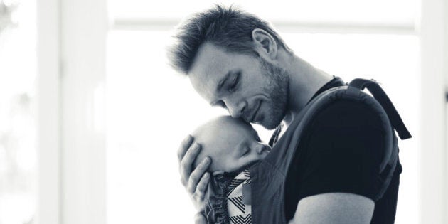 Young father holding his baby son in a sling in a living room. He is supporting his head with his hand and he is leaned to his head. He have his eyes closed and he is smiling. Little baby is sleeping. Black and white image.