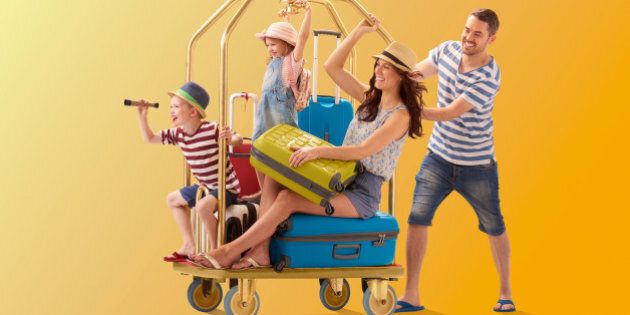 a young family climb aboard a luggage trolley and dad pushes them off to their holiday destination . They are all wearing warm weather clothing and holding their suitcases.