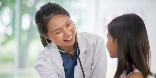 A little girl is at the doctors office for a check up. A doctor is listening to the child's heartbeat with at stethoscope.