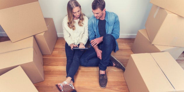 Young couple sitting in new house with packing boxes. They look like they have just moved in and they are very happy, smiling and talking. They are using a digital tablet, sitting on the floor in casual clothes. Copy space.