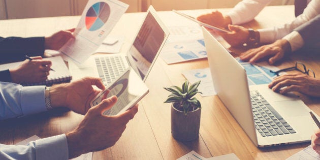 Group of people meeting with technology. They are around a conference table with digital tablets and laptops. There are marketing and strategy documents on the table