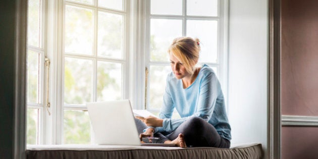 A full length photo of woman using laptop while holding documents at home. Young female is sitting on window sill. She is working from home.