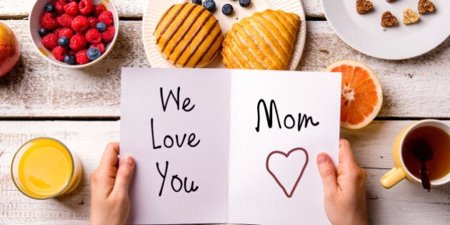 Mothers day composition. Hands of unrecognizable woman holding greeting card with We love you, Mom, text. Breakfast meal. Studio shot on wooden background.