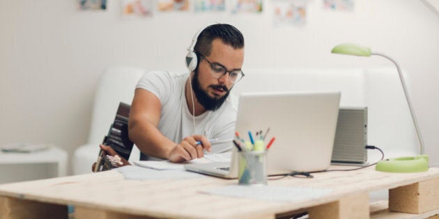 Man with acoustic guitar composing music for iStock and Getty stock library. Using laptop and writing lyrics or music notes. Wearing headphones around neck and typing on laptop.