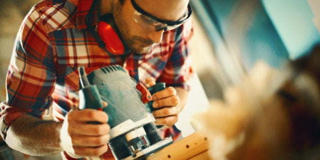 Closeup low angle shot of early 30;s man doing some carpentry work in a workshop. He's removing edges on a plank with a lunge router. Wearing protective glass and ear protectors. Tilt shot.