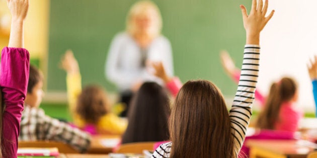 Rear view of large group of school children raising their hands ready to answer the question. Focus is on girl in striped shirt.