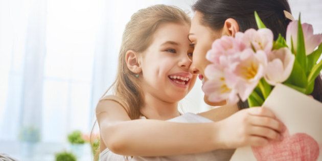 Happy mother's day! Child daughter congratulates mom and gives her flowers tulips and postcard. Mum and girl smiling and hugging. Family holiday and togetherness.
