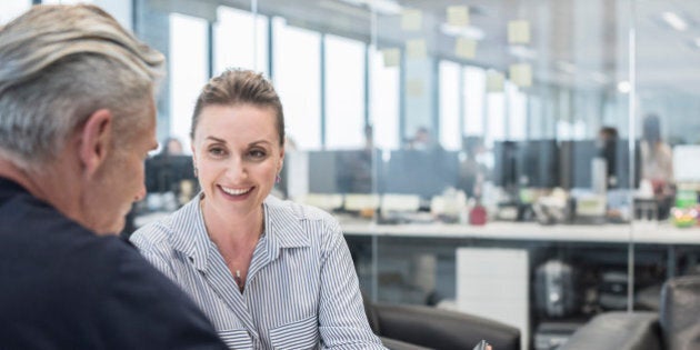 Two business people in informal meeting with digital tablet. Woman smiling towards man, explaining. Male client with female business woman in modern open plan office