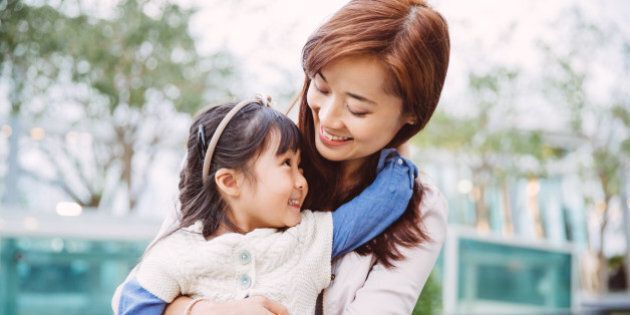 Lovely little daughter sitting on her pretty young mom's lap, both smiling joyfully face to face and looking into each others, while littler daughter reaching her hand back and hugging around mom's neck in the park