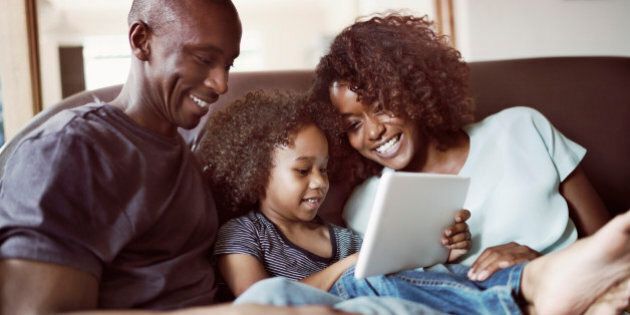 A photo of happy family sitting on sofa at home. Smiling parents are looking at son using digital tablet in living room. They are in casuals.