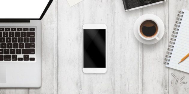 White smart phone with isolated screen for mockup on office desk. Laptop, camera, cup of coffee, paper, notepad, pencil on white table. Top view.