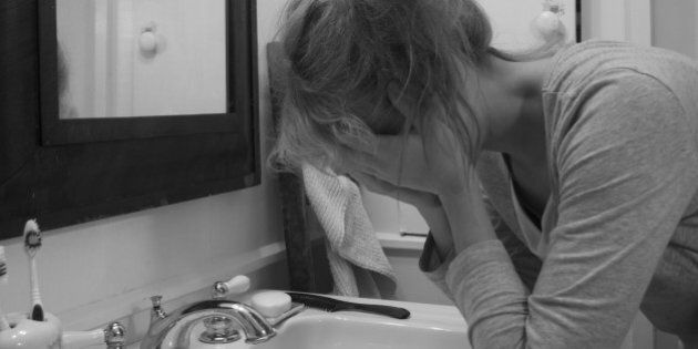 Black-and-white portrait of an upset woman slouching on a bathroom sink with her face in her hands, in front of a mirror.