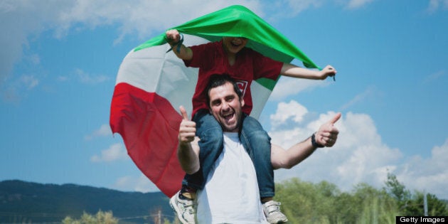 Son on dad's shoulders showing italian flag, they are walking and having fun outdoor.