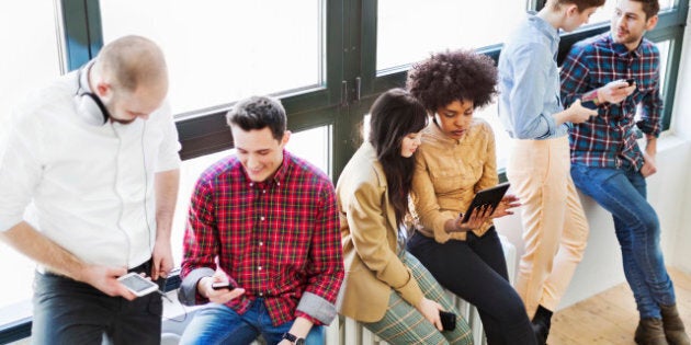 A group of young business people standing on a window talking and looking at their mobile devices. Backlit shot.