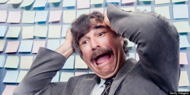 This is a horizontal, color photograph of an older mustached man sitting in front of a cubicle wall that is covered with sticky notes. He pulls at his hair and screams in frustration.