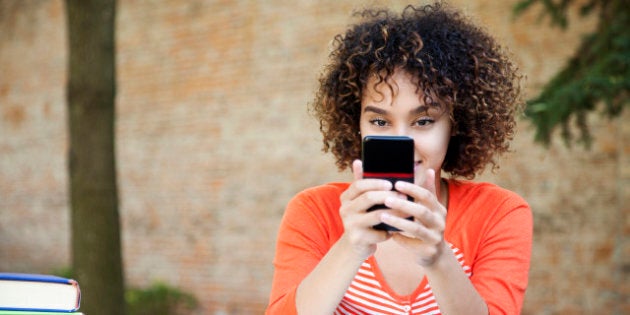 Ethnic female student with curly hair wearing wearing an orange shirt and texting with her smart phone