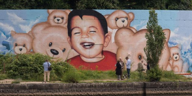 People stand in front of a new graffiti by artists Justus Becker and Oguz Sen depicting the drowned Syrian refugee boy Alan Kurdi (initially reported as Aylan Kurdi) at the harbor in Frankfurt am Main, Germany, on July 4, 2016.The artists created the new mural showing Alan Kurdi inmid of teddy bears after vandals had destroyed the former mural showing the Syrian toddler drowned. / AFP / dpa / Boris Roessler / RESTRICTED TO EDITORIAL USE - MANDATORY MENTION OF THE ARTIST UPON PUBLICATION - TO ILLUSTRATE THE EVENT AS SPECIFIED IN THE CAPTION---GERMANY OUT (Photo credit should read BORIS ROESSLER/AFP/Getty Images)