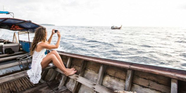 A woman in a white dress taking a photo with her iPhone while riding on a long tail boat to a tropical island in Thailand.