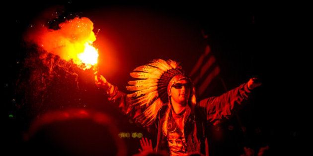 GLASTONBURY, UNITED KINGDOM - JULY 08: A man in the crowd wearing a Native American Headdress holding a lit flare as Jake Bug performs headlining The Other stage at the end of Day 2 of The Glastonbury Festival at Worthy Farm on July 8, 2014 in Glastonbury, England. (Photo by Ollie Millington/Redferns via Getty Images)