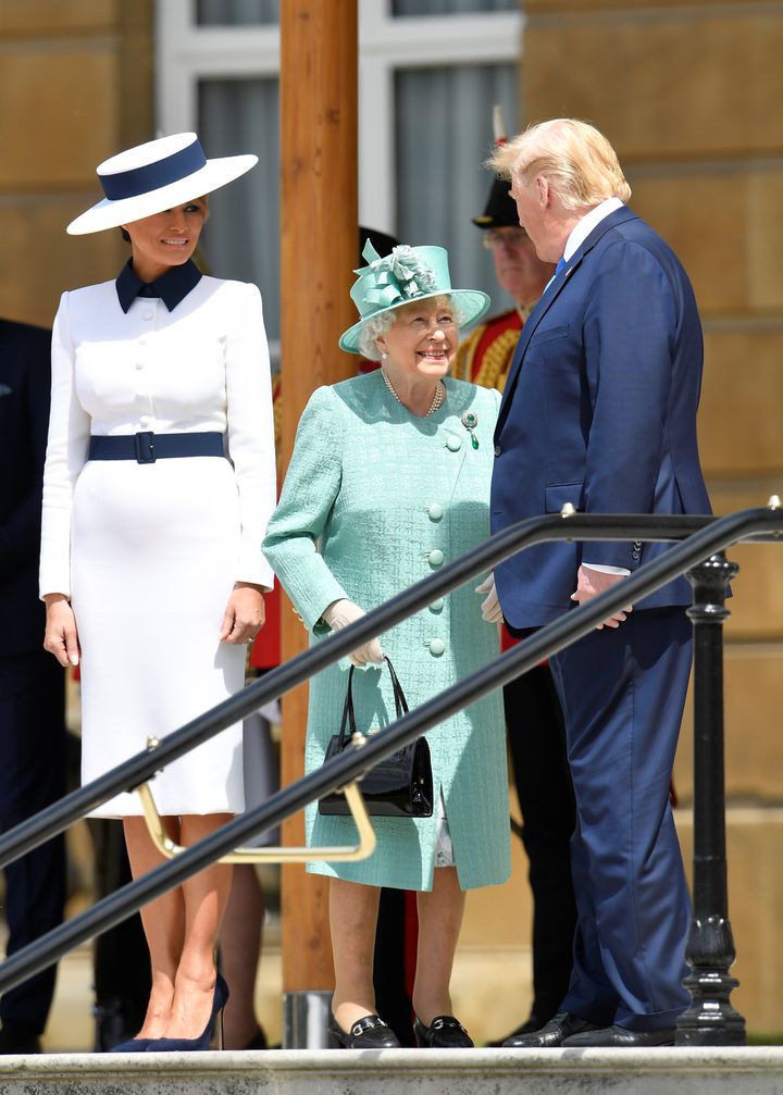 Melania Trump, the Queen, and Donald Trump, speak before observing a Guard of Honour at Buckingham Palace. The Queen is in a muted jade Stewart Parvin A- line coat and pleated dress in shades of grey, jade and dusky pink with a matching Rachel Trevor-Morgan hat.